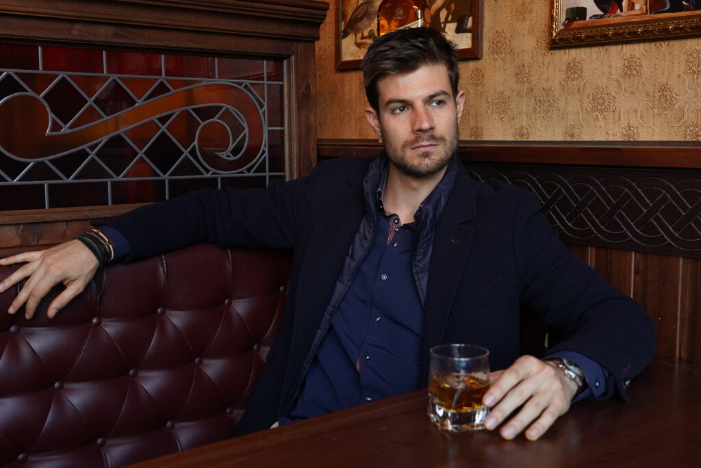 A young Hispanic male in a formal outfit sitting at a restaurant and drinking whiskey